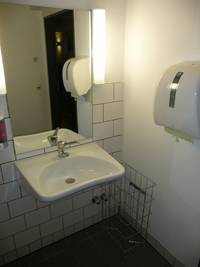 Auf dem Bild ist ein weißes Waschbecken mit silbernen Hahn zu sehen. Hinter dem Waschbecken hängt ein Spiegel an der Wand. Auf jeder Seite des Spiegels ist eine längliche Lampe zu sehen. Der Seifenspender sitzt links vom Spiegel, rechts vom Waschbecken steht ein Drahtgitter-Papierkorb. Rechts an der Wand hängt ein Papiertuchspender, dessen Spiegelbild im Spiegel zu sehen ist.